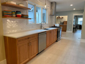 A bright kitchen remodel by H.E.L.P. featuring natural wood cabinets, a white countertop, herringbone tile backsplash, open shelves, and stainless appliances.