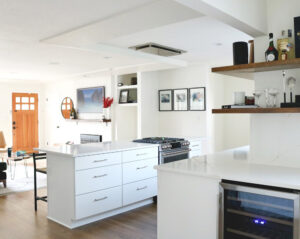 Alt text: After kitchen remodel by H.E.L.P. showcasing a bright open-concept layout with quartz countertops, white cabinetry, a central cooktop, floating shelves, and a built-in beverage fridge.