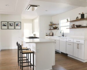 After kitchen remodel by H.E.L.P. featuring white cabinets, a large quartz waterfall island with seating, floating wood shelves, and warm wood flooring in an open layout.
