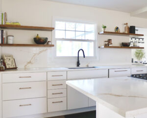 After kitchen remodel by H.E.L.P. featuring white cabinetry, a black faucet under a bright window, quartz countertops, and floating wood shelves for added storage and style.